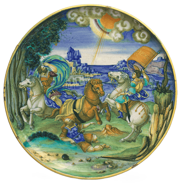 A shallow maiolica bowl with a painting of two men riding white horses and a third man who had fallen from his brown horse on the ground. In the sky above, the sun appears from behind clouds, with a ray directed toward the man on the ground. The scene depicts the biblical story of the conversion of Saul.