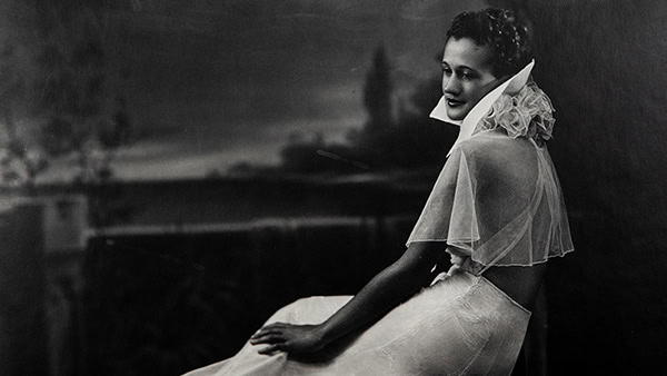 Black and white photograph by P. H. Polk titled Mildred Hansen Baker. 1930s era woman seated wearing a light colored high-collared formal gown gazing down over her left shoulder with her hand resting lightly in her lap.